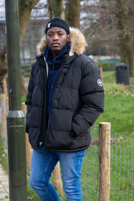 A young man standing outdoors on a paved pathway, wearing a black puffer jacket with a fur-lined hood, a dark blue hoodie underneath, and a navy knit beanie. His hands are in his jacket pockets, and he has a serious expression on his face. The background features a wire fence with wooden posts on the right, a metal pole on the left, and a grassy area with trees and shrubs beyond. There are also blurred elements of a park or garden setting, with a few bins visible in the distance, indicating an outdoor environment near a community or residential area. The scene is lit naturally, likely during the daytime with overcast weather, creating a neutral and muted tone that complements the winter clothing. This image aligns with the theme of outdoor rubbish removal or private waste management services, as indicated by the natural surroundings and the focus on outdoor environments, which are often associated with domestic or community clearance activities.