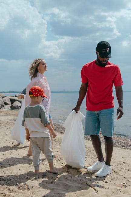 A young man wearing a black cap, red t-shirt, and light blue denim shorts is standing on a sandy beach, holding a large white plastic bag in his right hand while facing downward. To his left, a small child in a grey and white outfit with a red cap decorated with yellow polka dots is holding a similar white plastic bag, looking toward the ocean. Behind them, a woman with curly blonde hair, dressed in a light pink blouse, is gazing into the distance, also near the water's edge. The scene is set under a partly cloudy sky with patches of blue visible, and there are rocks lining the shoreline behind the group. The environment appears calm with a natural coastal backdrop, highlighting activities related to beach cleanup or casual waste collection, subtly reflecting the context of private waste handling and rubbish removal services such as those provided by Rubbish Collection Croydon.