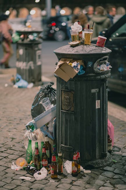 A black, cylindrical public trash bin with a domed lid, situated on a cobblestone pavement in an outdoor urban setting. The bin has various paper and plastic debris attached to its surface, and overflowing with discarded waste. Surrounding the base of the bin, there are several empty glass bottles, crumpled paper, and plastic containers, some of which are partially inside and others spilled onto the ground. On top of the bin, there are various used food and beverage items, including a paper food container, cups, and receipts. In the background, blurred figures of pedestrians and parked cars are visible, indicating a busy city street with natural daylight. The scene reflects typical urban rubbish that could be handled by private waste disposal services such as Rubbish Collection Croydon, emphasizing the importance of effective rubbish removal and on-site waste management.