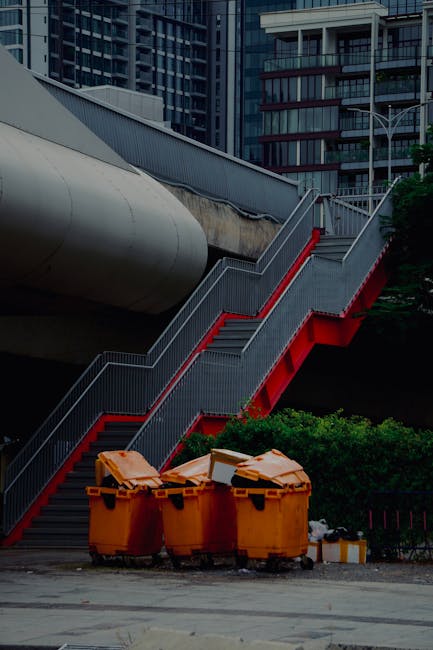 The image shows three large, orange wheeled waste bins placed on a paved surface in an urban setting, positioned in front of a green shrubbery. Behind the bins, there is a staircase with metal railings painted in gray and red accents, ascending alongside a concrete footbridge or walkway structure that is supported by a large, cylindrical concrete pipe running beneath it. The background features tall, modern office buildings with glass facades, indicating a city environment. The scene is captured under natural daylight, highlighting the textures of the metal staircase, the smooth surface of the waste bins, and the reflective glass of the surrounding buildings. This scene visually relates to private rubbish collection services, such as those provided by Rubbish Collection Croydon, and illustrates an ongoing process of waste disposal or collection in a commercial or residential urban area, emphasizing the importance of organized waste management and alternative disposal methods.