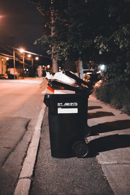 A middle-aged man with dark hair wearing a black T-shirt is seen discarding waste into a tall, cylindrical stainless steel waste bin on a paved outdoor pathway. He holds a large white plastic rubbish bag in his left hand and appears to be leaning forward to reach into the bin with his right hand. The man is using a wooden stick or pole to assist with placement of the waste. The surrounding environment features lush green trees and bushes, creating a park-like setting. The stainless steel bin has a smooth, reflective surface and an open top, positioned near a low stone wall that runs parallel to the pathway. The scene is well-lit with natural daylight, emphasizing the cleanliness and organized appearance of the rubbish disposal process. This image exemplifies private waste management or on-site rubbish collection, aligning with services offered by firms like Rubbish Collection Croydon engaged in independent rubbish removal efforts in outdoor urban environments.