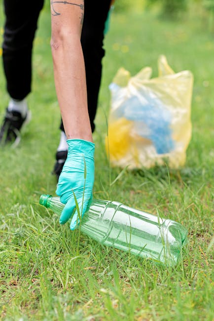 A person wearing a blue disposable glove is crouching down on a grassy area, holding a clear plastic bottle with a green cap and a ridged body, preparing to dispose of it. In the background, there is a yellow and white trash bag placed on the grass, partially filled with other plastic waste and household rubbish. The scene is outdoors, with a green, blurred natural environment suggesting a park or garden setting. The image reflects activities related to waste collection or environmental cleanup, pertinent to private rubbish removal services such as those provided by Rubbish Collection Croydon, emphasizing proper disposal of recyclable materials outside of local authority arrangements.