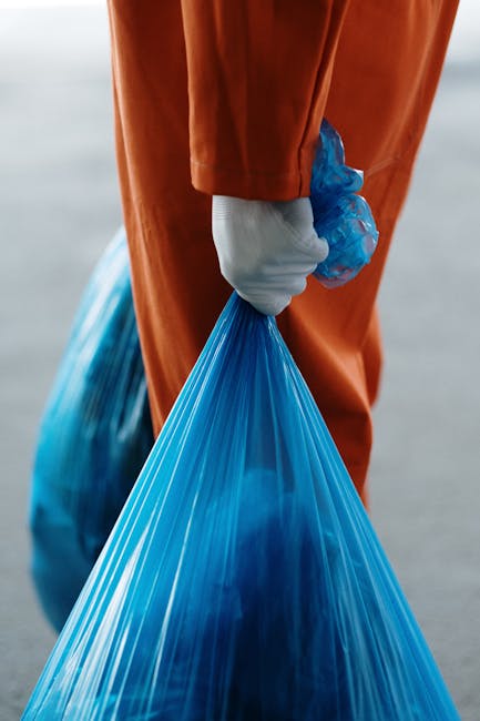 A person wearing a blue disposable glove is crouching down on a grassy area, holding a clear plastic bottle with a green cap and a ridged body, preparing to dispose of it. In the background, there is a yellow and white trash bag placed on the grass, partially filled with other plastic waste and household rubbish. The scene is outdoors, with a green, blurred natural environment suggesting a park or garden setting. The image reflects activities related to waste collection or environmental cleanup, pertinent to private rubbish removal services such as those provided by Rubbish Collection Croydon, emphasizing proper disposal of recyclable materials outside of local authority arrangements.