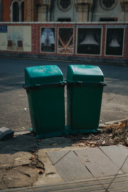 Two green wheeled rubbish bins with closed lids positioned side by side on a paved urban sidewalk near the edge of a road, with small leaves and debris on the ground around them. The bins are made of durable plastic, featuring textured surfaces and black handles for easy lifting. The scene is illuminated by natural daylight, with a blurred background of a brick and stucco building displaying decorative windows and a low brick wall. The bins appear to be for waste or recycling collection, likely used as part of local or private rubbish removal services, such as those provided by Rubbish Collection Croydon, to facilitate on-site waste disposal in residential or commercial areas.